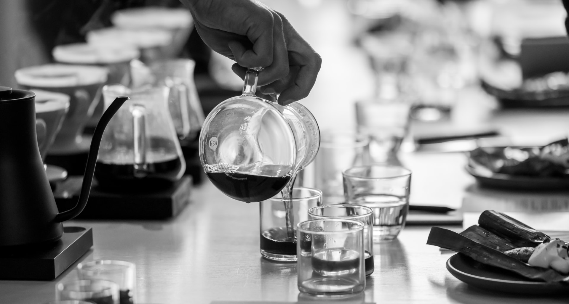 A black and white photograph of coffee being poured from a glass carafe into small cups on a set table